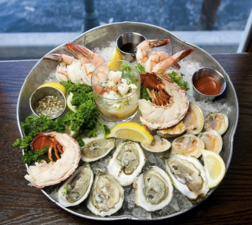 A seafood platter featuring shrimp, oysters, and a cocktail, garnished with lemon and herbs on ice.