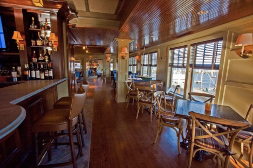 Interior of a cozy bar with wooden furniture, a long counter, and large windows overlooking a marina.