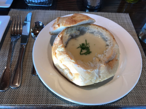 A bread bowl filled with creamy soup, garnished with herbs, on a white plate with utensils beside it.