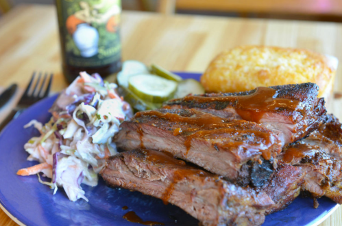 A plate of barbecue brisket with sauce, coleslaw, pickles, and a cornbread muffin on a wooden table.
