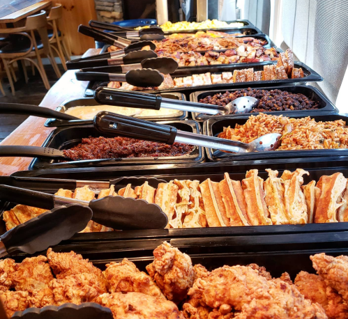 A buffet table with various dishes, including fried chicken, rice, beans, and waffles, all served in black trays.