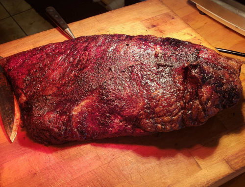 A large, smoked brisket resting on a wooden cutting board, with a knife beside it. The surface is dark and caramelized.