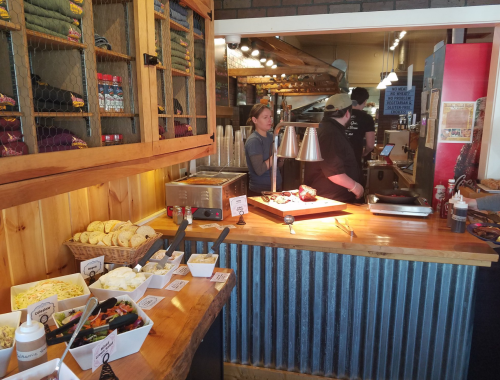 A cozy restaurant interior with a wooden counter, food displays, and staff preparing meals in a warm atmosphere.