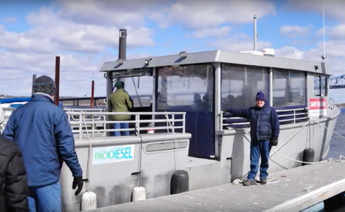 A group of people on a dock near a biodiesel boat, with one person waving at the camera. Cloudy sky in the background.