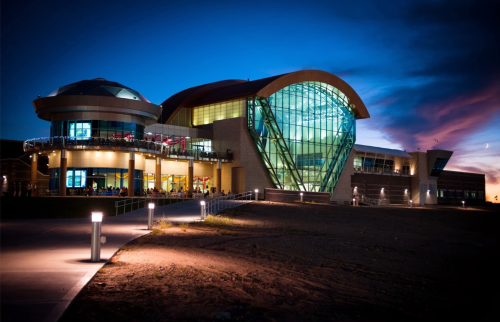 Modern building with a large glass facade, illuminated at dusk, surrounded by a pathway and a colorful sky.