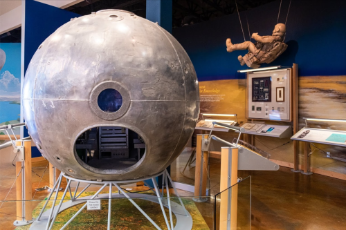 A metallic spacecraft model displayed in a museum, with an astronaut figure and informational panels in the background.