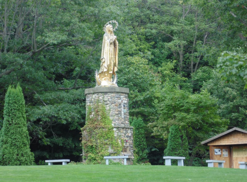 A golden statue on a stone pedestal surrounded by trees, with benches and a small wooden structure nearby.
