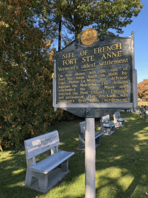 Historical marker for Fort Ste. Anne, Vermont's oldest settlement, with benches and trees in the background.