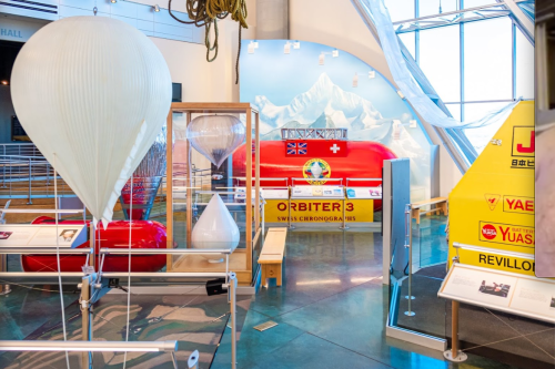 Exhibit featuring a red Swiss balloon, hourglass, and informational displays in a modern museum setting.