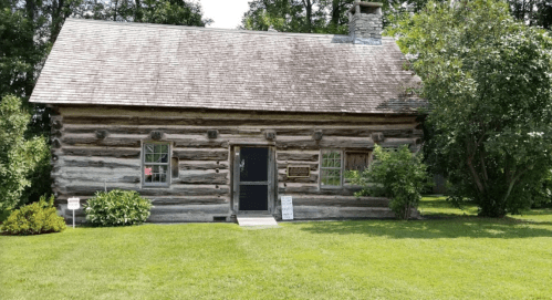 A rustic log cabin surrounded by greenery, featuring a stone chimney and a well-maintained lawn.