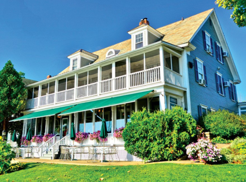 A charming blue house with a large porch, surrounded by colorful flowers and greenery under a clear blue sky.