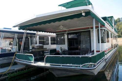 A houseboat docked at a marina, featuring a green awning, outdoor seating, and a grill on the deck.