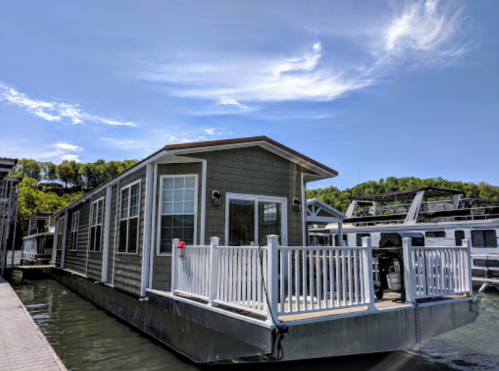 A houseboat docked at a marina, surrounded by water and greenery under a blue sky with scattered clouds.