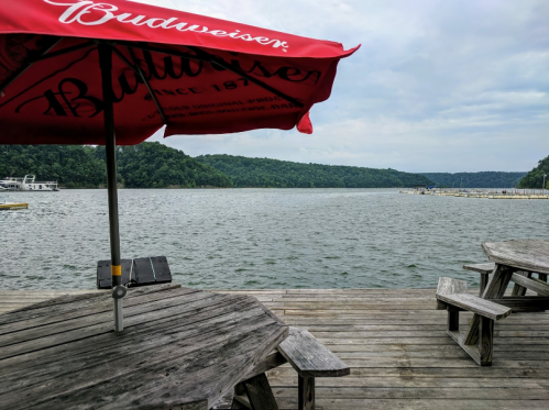 A wooden dock with picnic tables and a red umbrella overlooking a calm lake surrounded by green hills.