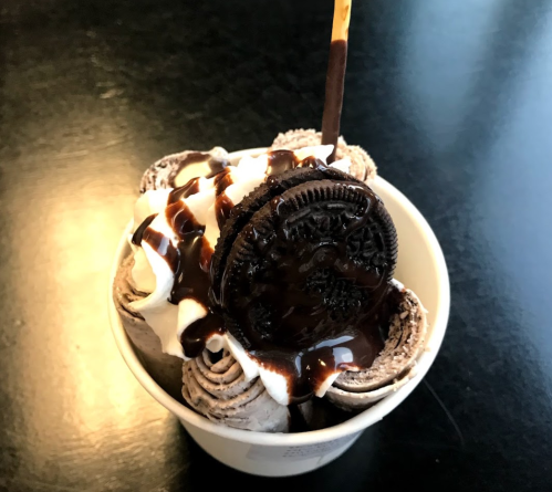 A cup of rolled ice cream topped with whipped cream, chocolate sauce, and an Oreo cookie.