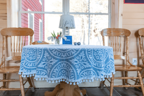 A cozy table set with a blue patterned tablecloth, a lamp, flowers, and a small decorative item by a sunny window.