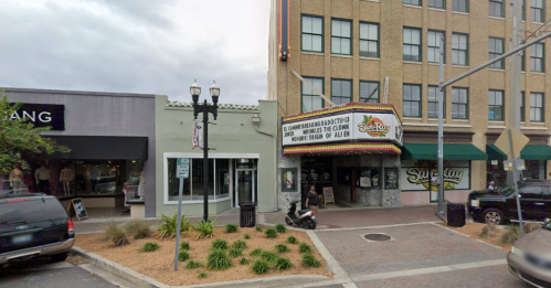 A street view of a theater with a marquee, flanked by shops and greenery along the sidewalk.