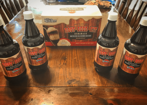 Four bottles of root beer sit on a wooden table, with a box of root beer extract in the background.