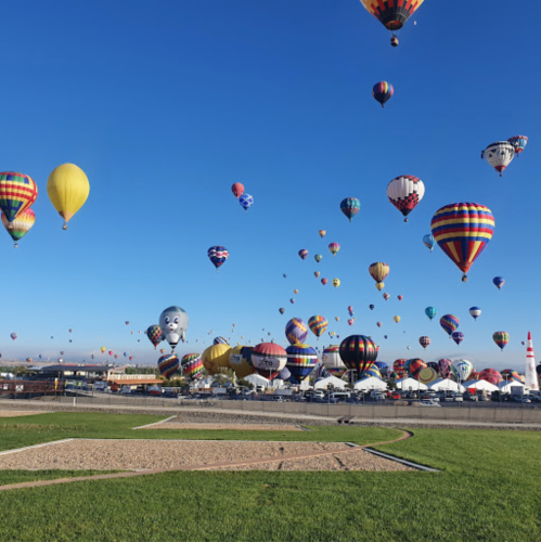 A vibrant sky filled with colorful hot air balloons at a festival, with a clear blue backdrop and a grassy field below.