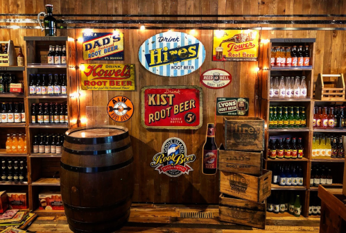 A rustic display of vintage root beer signs and bottles, with a wooden barrel and crates in a cozy setting.