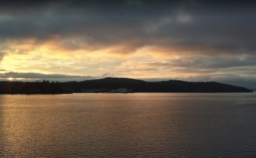 A serene sunset over a calm body of water, with silhouettes of hills and a ferry in the distance.