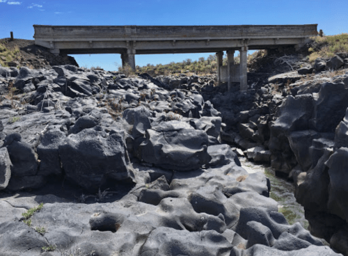 A concrete bridge spans a rocky landscape with dark volcanic rocks and a narrow waterway below.