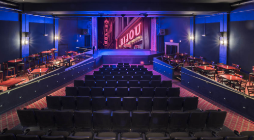 Interior of a theater with empty seating, a stage lit in purple, and tables set for an audience.