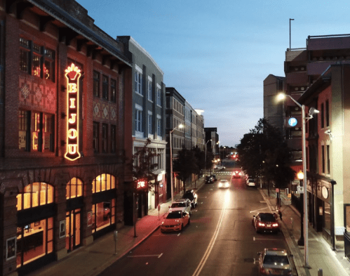 A city street at dusk, featuring the illuminated Bijou theater and parked cars along the road.