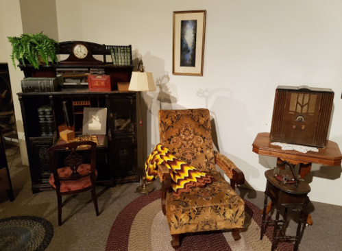 A vintage living room setup featuring an ornate chair, side tables, a lamp, and an antique radio against a neutral wall.