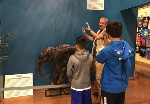 A guide explains a mounted baby mammoth exhibit to two children in a museum setting.