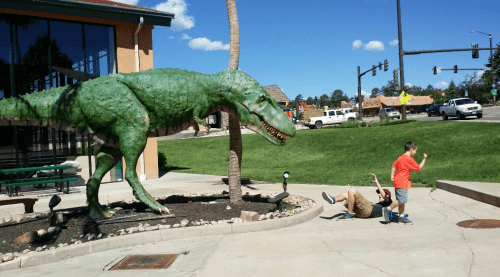 A child runs away from a large green dinosaur statue while another child falls on the ground nearby.