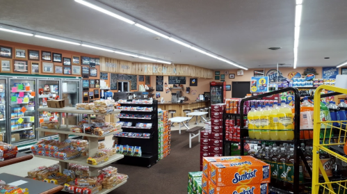 Interior of a convenience store with shelves of snacks and drinks, seating area, and a display of colorful signage.