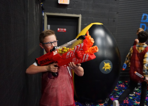 A child in glasses aims a red toy blaster in a colorful indoor play area, with a large black object in the background.