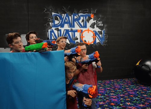 A group of kids in safety goggles aiming toy blasters behind a blue barrier in a colorful indoor play area.