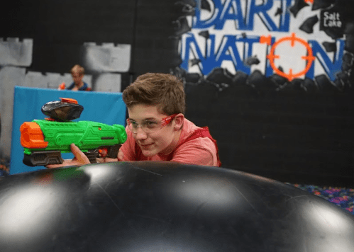 A boy aims a green toy blaster while crouching behind a large inflatable in a colorful indoor play area.