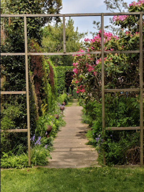 A garden path framed by a wooden trellis, lined with vibrant flowers and lush greenery on either side.