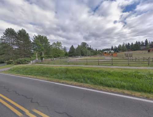 A rural scene featuring a grassy field, wooden fences, and a cloudy sky, with a road in the foreground.