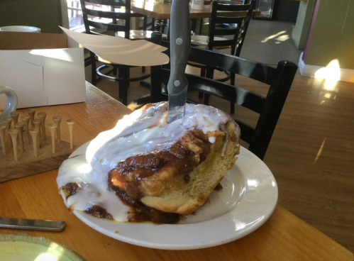 A large cinnamon roll with icing, topped with a knife, served on a plate in a cozy restaurant setting.