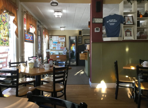 Cozy diner interior with wooden tables, black chairs, and colorful stained glass windows. Counter and decor visible.
