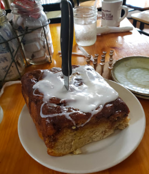 A large, frosted cinnamon roll on a plate with a knife inserted, set on a wooden table with coffee and condiments nearby.