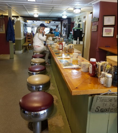 A diner interior with a long wooden counter, stools, and a woman standing at the bar, preparing food.