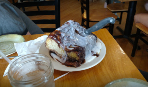 A large cinnamon roll with icing and a knife resting on top, served on a plate at a café.