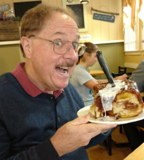 A man excitedly holds a large cinnamon roll topped with icing and a knife, smiling in a cozy restaurant setting.