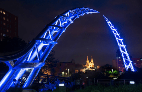 A brightly lit blue arch structure against a night sky, with buildings and a church in the background.
