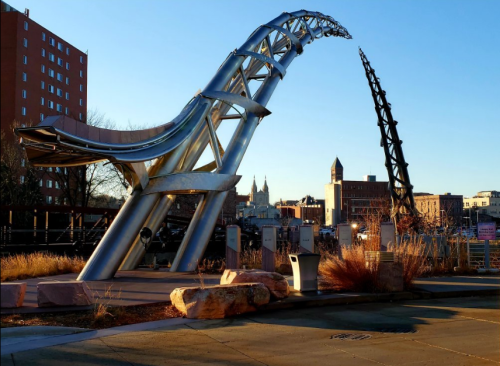 Two large, curved metallic sculptures rise against a clear blue sky, with buildings and a church in the background.