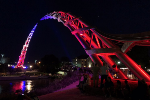 A brightly lit arch structure at night, with red and blue lights, overlooking a river and a crowd of people nearby.