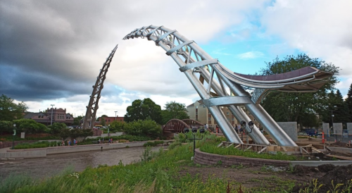 A modern metal sculpture with curved arches, set against a cloudy sky and greenery, near a river and buildings.
