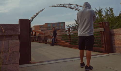 A person in a hoodie stands on a bridge, while another person in a uniform watches from a distance.