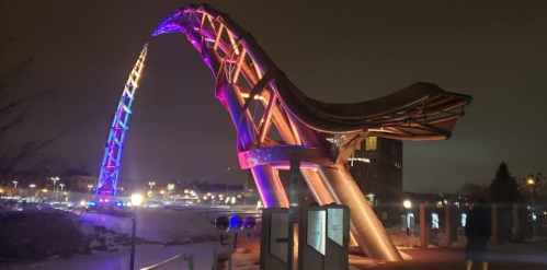 A colorful, illuminated sculpture resembling a wave or arch at night, surrounded by a snowy landscape.