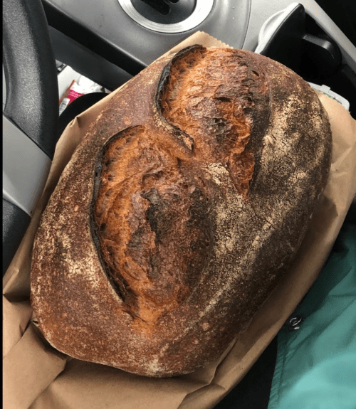 A freshly baked loaf of bread resting on brown paper, placed on a car seat next to a steering wheel.